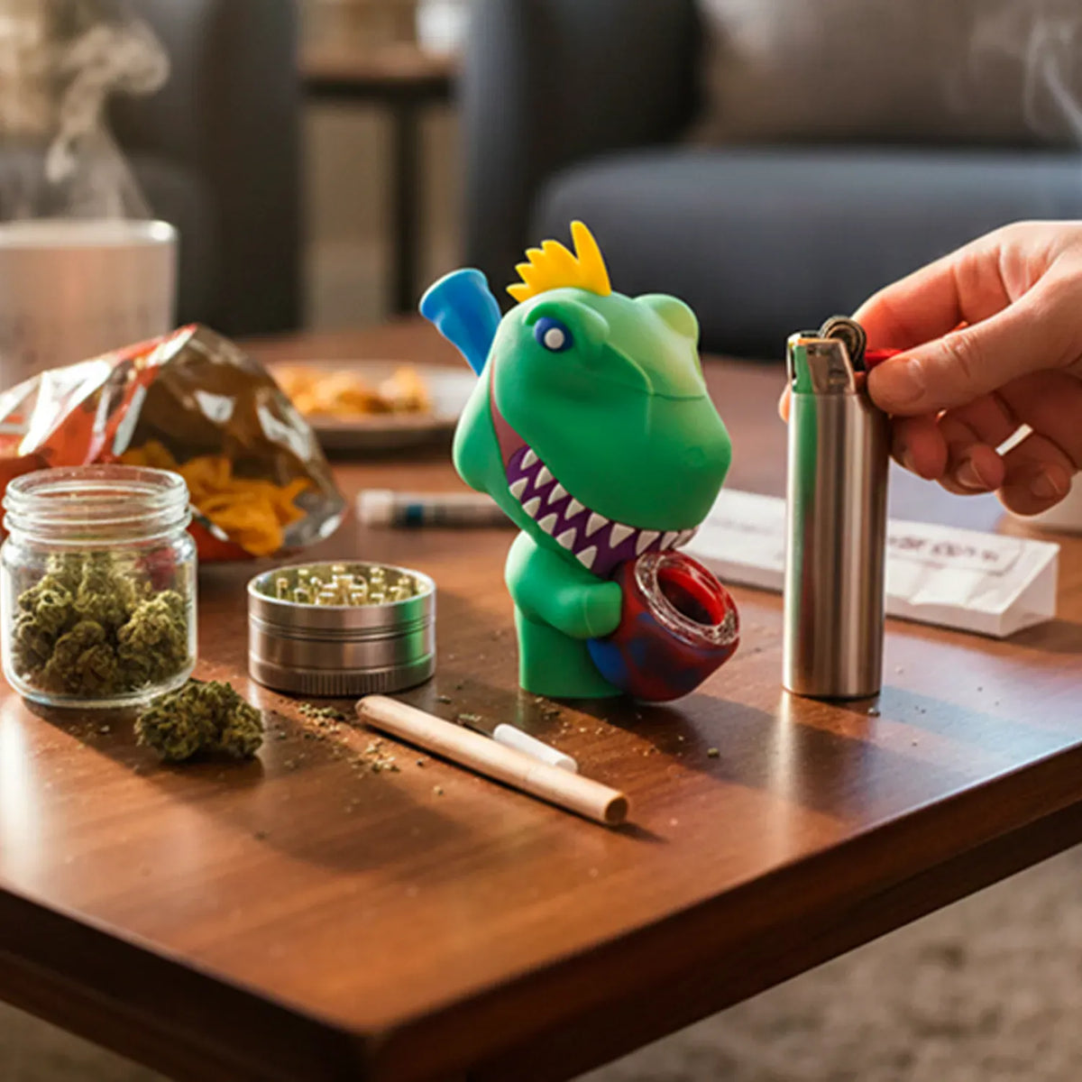 Tabletop view of a green Dino Silipipe dinosaur pipe with a red bowl held beside a lighter amid rolling papers, grinder, and jar of herbs.